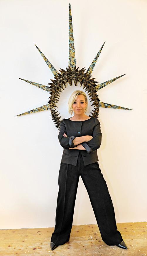 Sabine Wiedenhofer in dunklem Anzug vor einer strahlenkranzförmigen Wandarbeit